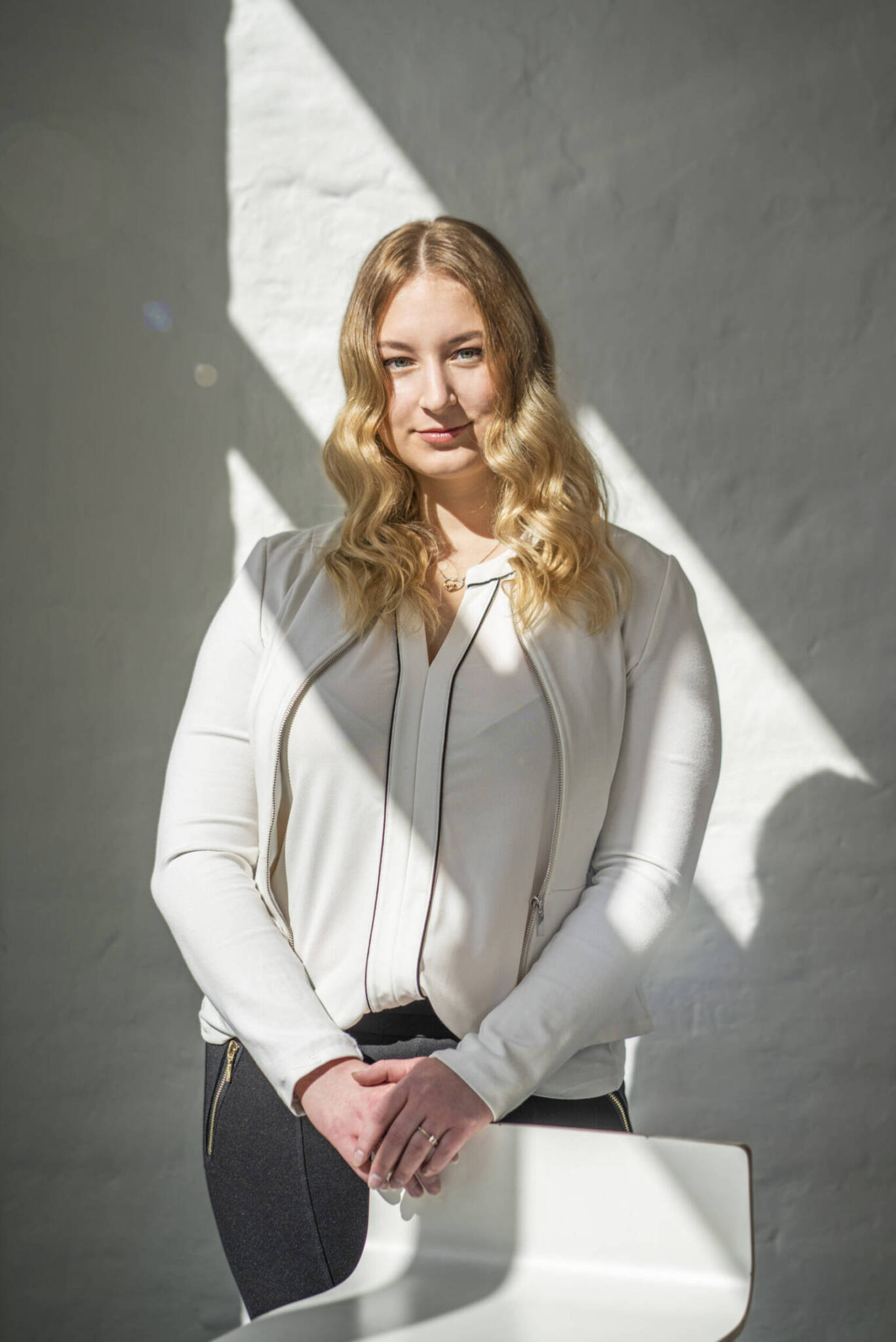 Mitarbeiterin steht im Sonnenlicht vor weißer Wand, Blick in die Kamera – professionelles Businessportrait