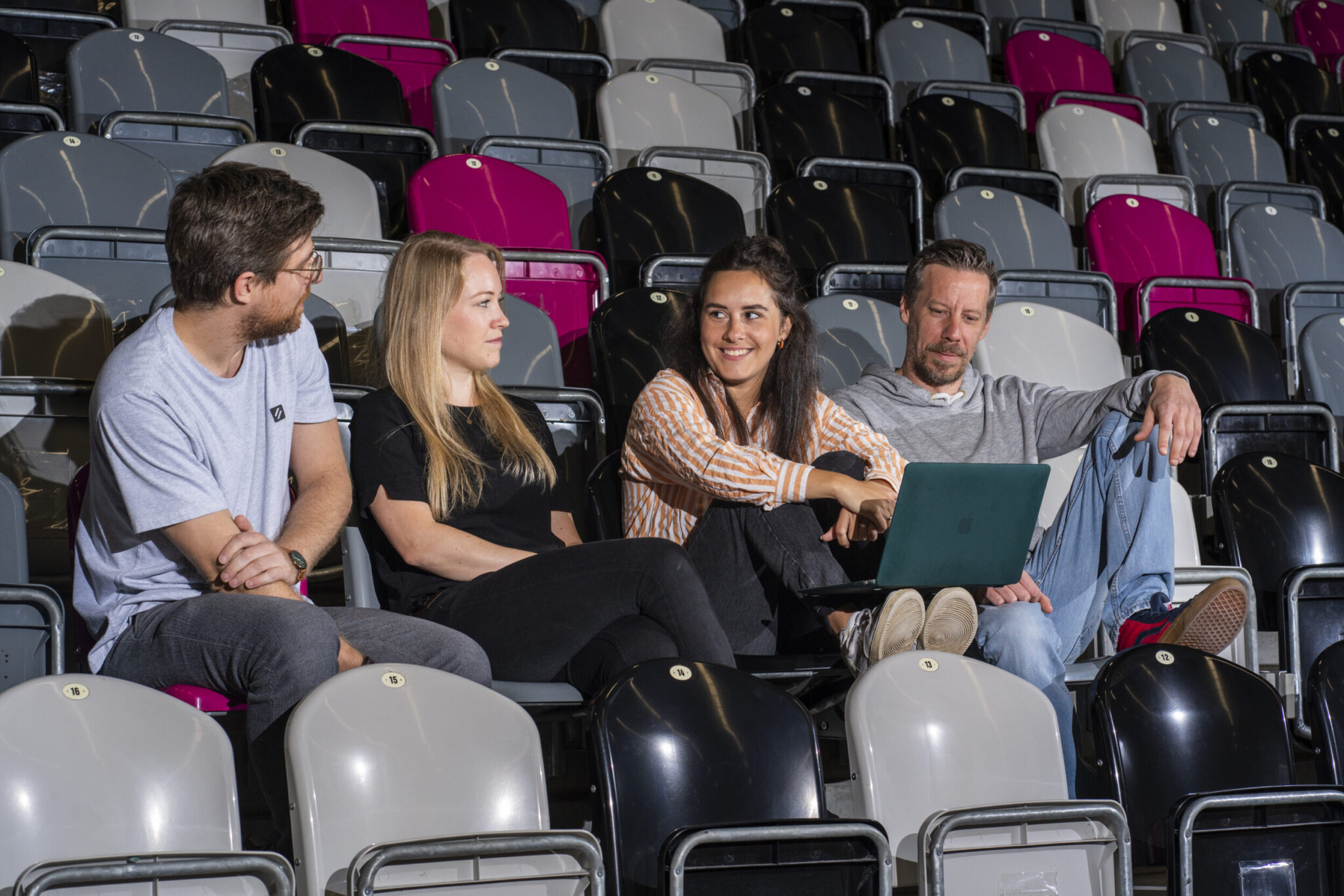 Vier Mitarbeitende sitzen auf Stadiontribüne, diskutieren entspannt mit Laptop – Vorbereitung für Unternehmensfotoshooting